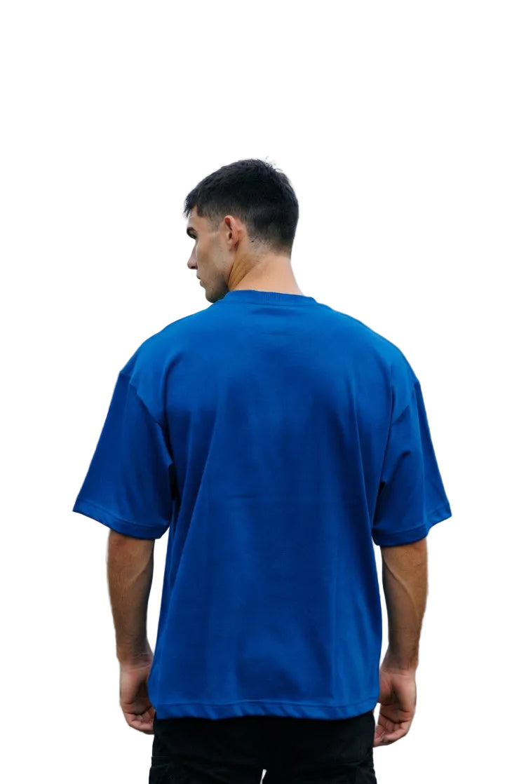 Man wearing a blue t-shirt standing on a residential street with trees and houses in the background.